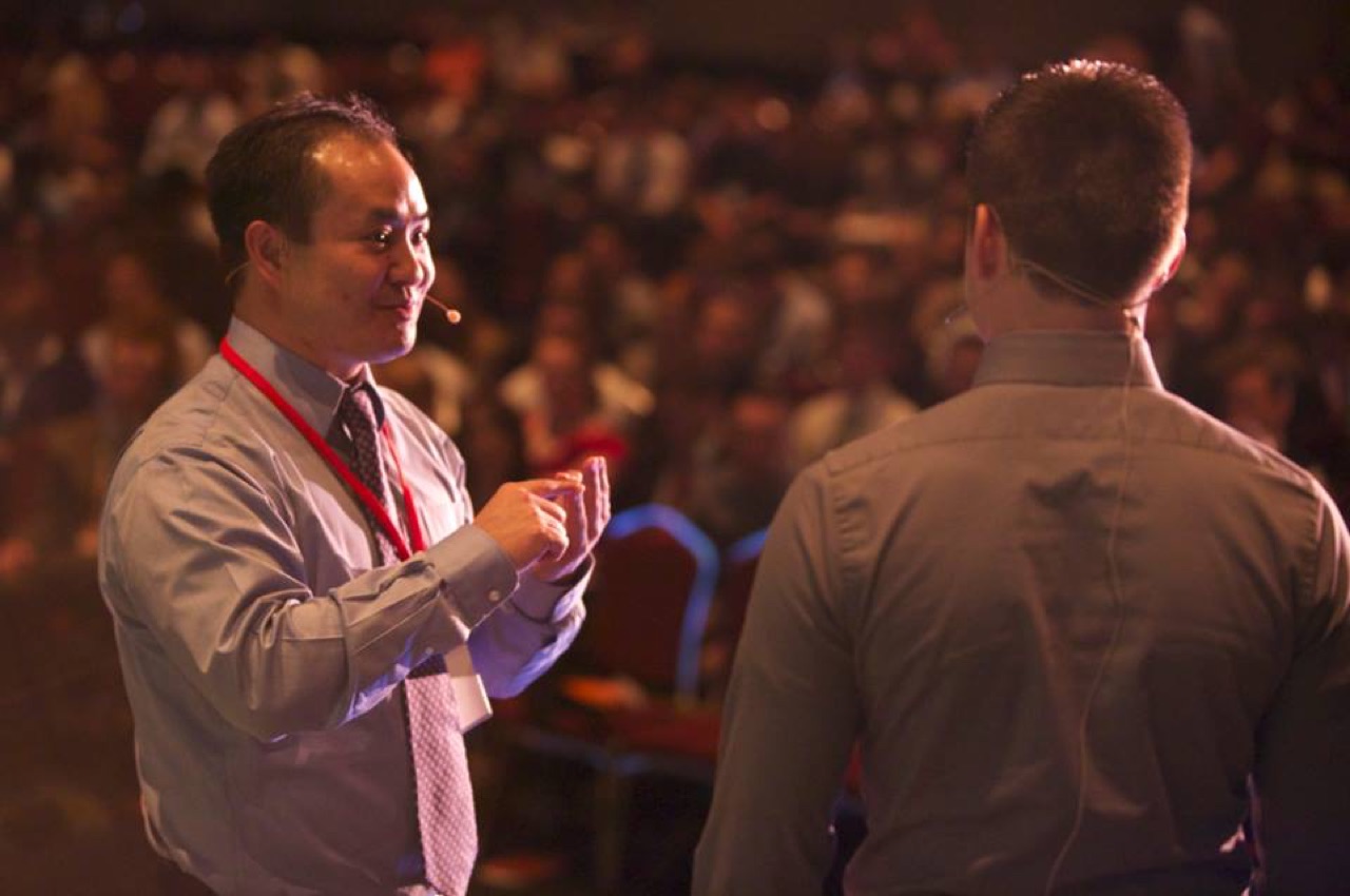 Dennis Yu coaching a young speaker on stage in front of a full house